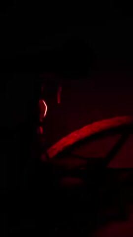 In a dark environment, a bicycle wheel is illuminated by red and sometimes purple light. A person wearing headphones or ear protection is partially visible to the left of the wheel. The focus is on the wheel and the changing color of the light.