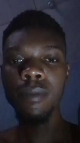 A close-up, head-on shot depicts a young, dark-skinned man with short hair and a trimmed beard. He is shirtless and appears to be indoors under dim lighting. The background is a blurry wall with a pattern. In the video, he places a bite of food, appearing to be a light-colored doughy item, into his mouth and chews. After swallowing, he inserts a dark-colored finger food into his mouth.