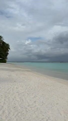 A wide shot shows a serene beach scene in the Maldives. The foreground is dominated by pristine white sand, gently sloping towards the turquoise ocean. The water is calm and clear, with shallow areas transitioning to deeper, darker blue further out. In the distance, a dark, ominous line of storm clouds stretches across the horizon, contrasting with a small patch of lighter sky.

The main subject is a young girl wearing a light-colored bikini. She is walking along the water's edge, her feet occasionally touching the shallow waves. Her body language suggests a relaxed and contemplative mood. The overall atmosphere is one of natural beauty with a hint of impending weather change. The time of day appears to be late afternoon or early evening, given the diffused light and the dramatic cloud formations. There is no visible text in the media.