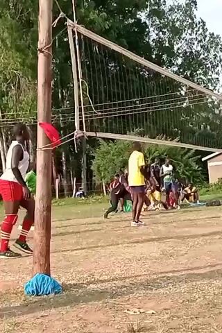 The video shows a group of people playing volleyball on a dirt court. There is a net separating the two sides, and a small building in the background. The players are wearing various athletic clothes. One of the players in a white shirt and black leggings jumps high to spike the ball over the net. Some people sit on the sidelines watching the game. The weather appears to be partly cloudy.