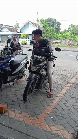 A young man in a gray sweater and light beige pants stands next to a black Honda motorcycle parked on a brick sidewalk near a road. He is fiddling with something on the motorcycle's dashboard. A young woman wearing a hijab and gray pants stands next to him, looking at the same area. There's another parked blue and white Honda motorcycle to their left. In the background, across the road, are houses and greenery. A person in a reflective vest is visible further away on the left side of the frame. The scene appears to be outdoors during the day.