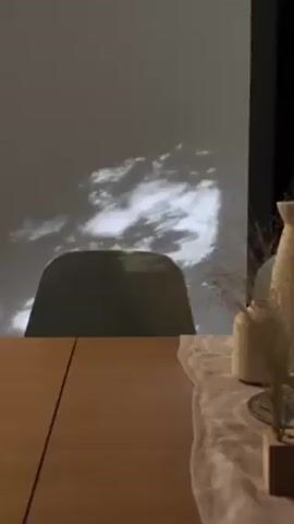 Here is a concise description of the video:

The video shows a room with sunlight filtering through trees, casting shadows onto a white wall. A chair sits in front of the wall, and a wooden table is visible in the foreground. There are decorative items on the table, including vases and dried plants.  The scene is peaceful and serene, emphasizing natural light and minimalist decor.