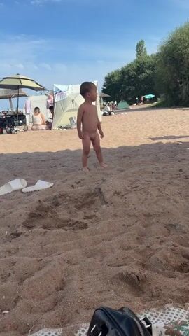 The video captures a sunny day at a beach in Choktal, Kyrgyzstan. The primary subject is a young child, likely a toddler, standing unclothed on the sandy shore. The child is facing slightly to the left, with their gaze directed downwards.

In the background, several tents and beach umbrellas are set up, indicating a populated area. A person, possibly an adult, is seated under one of the umbrellas. Further in the background, more people can be seen milling about, enjoying the beach. The sky is blue with scattered white clouds, suggesting pleasant weather. The scene is illuminated by bright sunlight, casting shadows on the sand.

A pair of white flip-flops are visible in the foreground on the sand, and a black bag rests on a patterned mat. To the right of the child, a person wearing a grey hoodie and shorts is partially visible, standing and looking downwards. The overall atmosphere appears relaxed and summery. There is no visible text in the video.