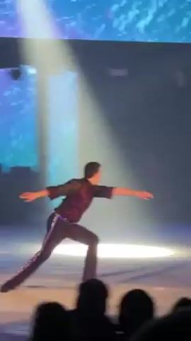 The video shows an ice skater on a stage, illuminated by a spotlight, performing a move on one knee with arms spread wide. The skater is wearing black pants, a gray shirt with a red collar, and ice skates. Behind the skater is a large dark screen and a smaller screen showing blue and white graphics. An audience is visible at the bottom of the frame. The audience cheers after the skater finishes the move.