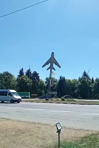A statue of a jet is visible against a clear blue sky. Trees surround the statue. A white van drives past the statue on the left side of the road. A woman is standing to the right of the statue.