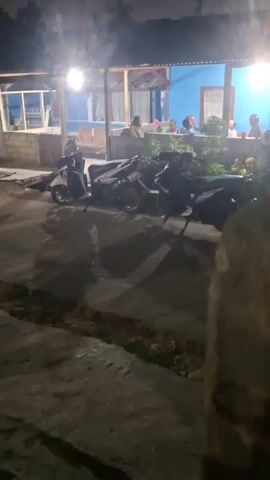 The video captures a dimly lit outdoor scene at night, likely a roadside eatery or a small cafe. The foreground is dominated by a cracked, dark concrete surface with some visible debris. Several motorcycles are parked in a row on the concrete, their shapes silhouetted against the faint ambient light. Beyond the motorcycles, there is a structure with an open-air seating area. Inside this area, several people are seated at tables, illuminated by bright overhead lights. The back wall of the structure is painted a vibrant blue. The overall atmosphere is casual and suggests a common gathering place.