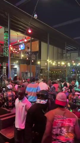 The video shows a bustling outdoor bar area, possibly at night, filled with people. Many are standing, seated at tables, and some are dancing. The crowd is diverse in age and attire, with some wearing hats, casual clothing, and colorful shirts. There’s a band or DJ performing on a raised platform to the right. String lights and various lamps illuminate the space, which has a multi-level structure, and there are drinks on the tables. The atmosphere appears lively and celebratory.