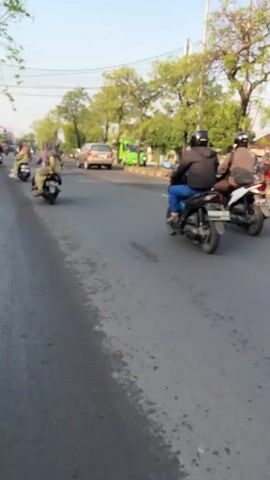The video shows a street scene. In the foreground, a gray asphalt road with some wet patches. In the middle ground, two people on a motorcycle are riding away from the camera, with the passenger wearing a dark hoodie. Behind them, another motorcycle is also moving away. Further back, there are cars, a green truck, and trees lining the road. In the background, there are some buildings and more trees. The sky is clear and a light blue, suggesting it is daytime.