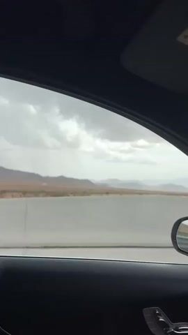 The video is filmed from inside a car, looking out the passenger window. The view shows a highway with a concrete barrier. Beyond the barrier, there are mountains in the distance under a cloudy sky. The interior of the car is visible, including the dashboard, a side mirror, and a hand on the steering wheel. The car appears to be in motion.