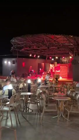 A band is playing on a stage at night. There are tables and chairs in front of the stage where people are seated. The stage is lit with red and green lights.