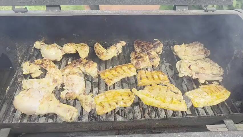 The video shows an overhead view of a grill with various pieces of chicken cooking. The chicken pieces include legs, thighs, wings, and breasts. Some pieces have grill marks on them, and there is visible smoke rising from the grill. The grill is filled with hot coals, and the chicken is arranged on the grates. The background is out of focus, but appears to be outdoors with greenery.
