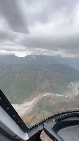 The video captures an aerial view from inside an aircraft, likely a helicopter, flying over a mountainous region under an overcast sky. The foreground shows the aircraft's cockpit, including a portion of the windshield with raindrops or mist, a section of the dashboard, and part of the internal structure on the left. Faint text "JETA-4" and numbers like "90 90 92 97" are visible on a panel on the right side of the dashboard.

The landscape features rugged, green mountains, with some areas appearing cultivated or terraced, and scattered small settlements or buildings visible on the slopes. A wide, sediment-rich river, light brown in color, winds through the valley below, with sandy or rocky banks. The sky is heavily clouded with thick gray clouds, partially obscuring the peaks of distant mountains. The weather appears to be rainy or recently rainy, with poor visibility due to the low cloud ceiling and moisture on the windshield. The scene is consistent with the provided context of Thulachhap, Nepal, showcasing a typical Himalayan foothill environment.