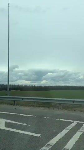 The video captures a view from a moving vehicle, likely a car, traveling along a paved road. In the foreground, the dark asphalt of the road is visible, with white road markings on its edge. A gray metal guardrail runs horizontally across the mid-ground, acting as a barrier. Beyond the guardrail, a wide expanse of green field stretches towards the horizon. In the distance, a dense line of dark trees, possibly a forest, forms a natural boundary. The sky above is heavily overcast with thick, gray clouds, suggesting a cloudy day. Around the 0:01 mark, a tall, slender streetlamp with two light fixtures appears on the right side of the frame, indicating the road might be part of a developed area or leading to one. No people are visible, and there is no discernible text. The scene depicts a rural or semi-rural landscape, consistent with the provided location context of Khutor, Russia, under overcast daylight conditions.