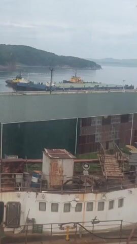 This is a static, vertical video of a dockside scene. In the foreground, the upper deck of a white ship is visible, with several windows along its side. A rusted metal structure, possibly a small shed or storage unit, is on the deck, along with a blue barrel. A wooden staircase leads up to this structure. Behind the ship, a large, dark green, gabled building dominates the middle ground, with corrugated metal panels visible on its side. Beyond the building, two tugboats are docked in a body of water, with a long, flat barge positioned between them. In the background, a treed, hilly coastline is visible under a cloudy sky. The overall impression is of a somewhat industrial and weathered maritime environment. No people are visible, and no actions are taking place beyond the implied presence of the docked vessels.