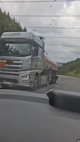 A silver tanker truck is driving on a paved road. The truck has a number plate that is difficult to read due to its angle. The truck has the words "FAW" written on it. The tanker itself is red and white. The truck is driving on a road that curves to the right. The road is lined with trees on both sides. The sky is overcast. The video appears to be shot from inside another vehicle.