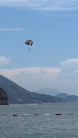 A video shows a parasailer in a colorful parachute high in the sky over a body of water. The parasailer appears to be suspended by a tow rope connected to a boat that is visible in the distance, moving across the water. The water is calm and there are buoys in the foreground that appear to be marking a boundary. In the background, there are mountains with some vegetation. The sky is blue with some scattered clouds. The overall scene depicts a recreational activity on a clear day.