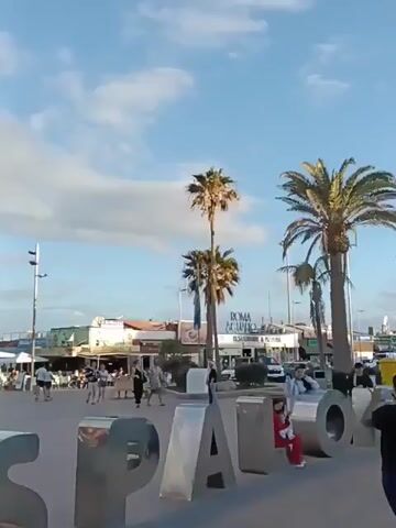 The video shows a view of the beach promenade in Santa Pola, Spain. In the foreground, there is a large, silver "SP" sculpture. People are walking along the paved promenade, and some are seated at outdoor tables with white umbrellas. Palm trees are visible, and the sky is blue with some white clouds. The background includes buildings with restaurants and shops.
