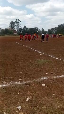 The video shows a soccer field with a red dirt surface. A group of people in red soccer uniforms is standing near a white line, presumably the edge of the field. Another group of people, some wearing yellow jerseys, are further away on the field. Several individuals in casual clothing are also visible near the red-clad players. The sky is partially cloudy, and some trees are visible in the background, suggesting an urban or suburban setting. There's white litter strewn on the foreground. The focus is on the players near the white line, with some activity or discussion appearing to be taking place. The camera appears to be hand-held.