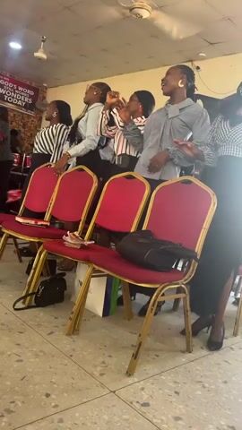 Here is a description of the video:

The video captures a group of women singing and clapping in what appears to be a church or a similar venue. They are standing behind a row of red and gold chairs on which purses and envelopes are placed. The women are dressed in stylish attire.  A band can be heard providing music. A banner is visible in the background.