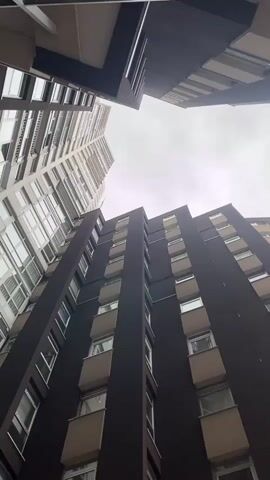The camera angle is low, looking up at multiple tall buildings against a pale, overcast sky. The buildings are a mix of dark and light tones, with many windows visible on each floor. The architectural style appears modern with clean lines. There's no people visible in the shot. The camera is still.