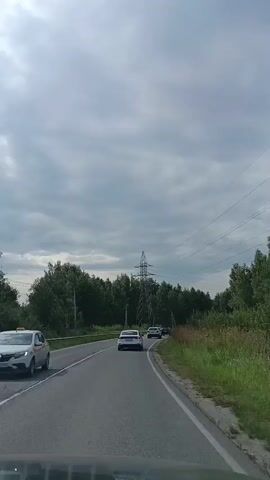 The video shows a view from inside a vehicle traveling on a paved road. The road is lined with trees on both sides, and tall power lines with multiple wires stretch across the sky. Several cars are visible on the road ahead, also traveling in the same direction. The sky is overcast with gray clouds. The timestamp indicates the video is approximately 5 seconds long.