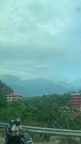 The video shows a landscape of rolling hills covered with lush green trees and buildings scattered among them. In the background, snow-capped mountains rise into a cloudy sky. The view is observed from a moving vehicle, as evidenced by the roadside barrier in the lower-left corner and the changing perspective of the buildings and surroundings. An electricity pole briefly enters the frame. The overall atmosphere of the scene is calm and serene, with the natural beauty of the mountains contrasting with the man-made structures in the foreground.