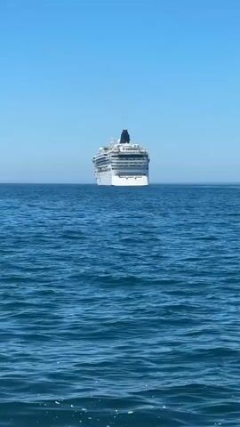 A large white cruise ship is visible on the horizon under a clear blue sky. The camera is positioned on a boat moving through calm blue water, creating a wake. Another smaller boat with orange accents is also in the distance, to the right of the cruise ship.