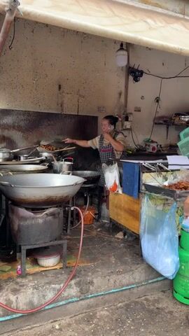 A woman with her hair pulled back in a bun, wearing an apron over a patterned shirt, stands in what appears to be an outdoor food stall. She is holding a mobile phone to her right ear with her right hand and using her left hand to stir food in a wok. The wok is positioned over a large, dark-colored stove. To her left, a large, silver pot is visible. Behind her, a wooden counter is filled with various items, including plastic bags, paper, and containers. A green gas cylinder is visible to the right of the stall. The environment is somewhat cluttered and appears to be outdoors, with a light-colored awning overhead.