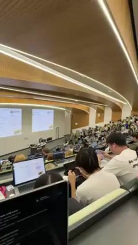 The video shows a wide view of a college lecture hall filled with students. A professor stands at the front of the room lecturing, with two projection screens displaying information. Many students are sitting at desks equipped with laptops or tablets. In the foreground, a student works on a laptop while another student behind them uses a smartphone. The lecture hall features a modern design with curved rows of seats, wood paneling on the ceiling, and bright, uniform lighting.