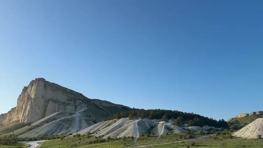 The video shows a panoramic view of a mountainous landscape under a clear blue sky. The camera pans from left to right, starting with a striking white rock formation with vertical cliffs. The landscape includes rolling hills with patches of green grass and scattered trees, some of which appear to be conifers. The ground appears to be a mix of exposed white rock and green vegetation. A dirt path is visible winding up one of the hills. The panoramic view ends on a plateau-like mountain with a rocky edge and green vegetation on top. The overall impression is of a scenic, natural environment.
