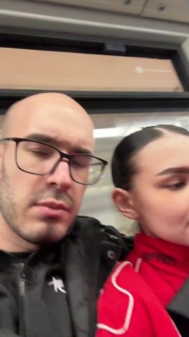 A woman wearing a red jacket in a subway car takes a selfie video. She holds up her phone which shows the selfie video she is taking. A debit card is held in her hand. A man with a bald head and glasses appears at the end of the video.