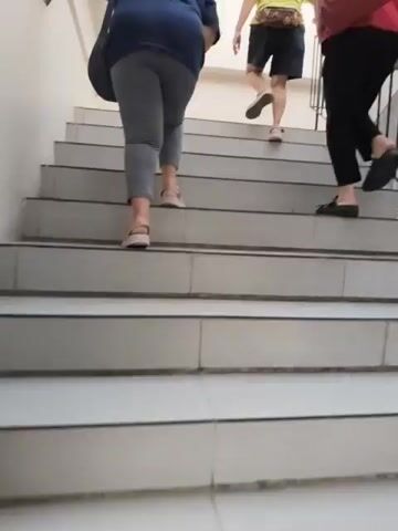 Three individuals are walking up a staircase. The first person, on the left, is wearing a blue shirt and gray leggings, and is holding onto the wall. The second person, in the middle, is wearing a yellow shirt and black shorts. The third person, on the right, is wearing a red shirt and black pants, and is carrying a red bag. The staircase is made of concrete and tile. The environment appears to be indoors.