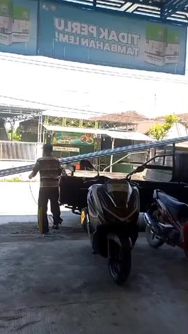 A man wearing a brown and tan striped shirt, dark pants, and a baseball cap is pulling a long metal pole from the back of a black pickup truck. The man is holding a rope that is attached to the pole, and he is walking backwards, dragging the pole. The pickup truck is parked under a covered area. A motorcycle is parked to the right of the pickup truck. There is a large sign in the background that says "TIDAK PERLU TAMBAHAN LEM!". The scene appears to be an outdoor storage or work area.