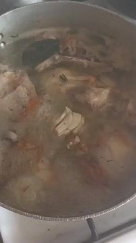 A close-up shot captures pieces of chicken and vegetables simmering in a pot of broth on a stovetop. The pot, made of a silvery metal, is filled with bubbling liquid, indicating that the contents are being cooked. Visible ingredients include chunks of chicken, possibly with bones, some chopped carrots providing an orange hue, and a few bay leaves for seasoning. The lighting suggests an indoor setting, possibly a kitchen, with no clear indication of the time of day or weather. The focus is entirely on the food preparation, with no people or discernible emotions present. The scene is static, with only the movement of the boiling liquid. There is no visible text in the image. The location context provided is Pochaiv, Ukraine.