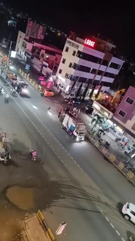 The video shows a night scene on a street with several vehicles and pedestrians. A large truck with a decorated trailer, possibly for a procession or event, is moving along the road. There are also motorcycles and a few cars on the street. Buildings with illuminated signs, including one that reads "LODGE," line the sides of the road. The street appears wet, possibly from recent rain, and there are puddles visible. Some people are standing on the side of the road, and others are on the vehicles. The overall atmosphere seems to be one of activity and movement.
