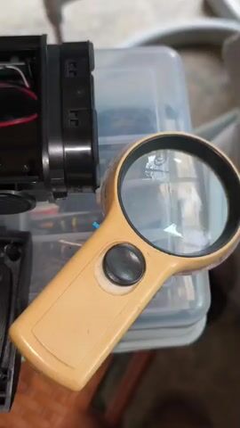 The video shows a close-up view of a disassembled speaker. A screwdriver is used to work on the internal components of the speaker. In the foreground, a magnifying glass rests on a clear plastic container. The speaker's black casing is open, revealing a green battery pack and circuit board. Two circular speakers are visible in the lower part of the frame. The overall environment appears to be a workbench or a cluttered area, with various tools and parts scattered around. The lighting is adequate for viewing the details of the speaker's internals.