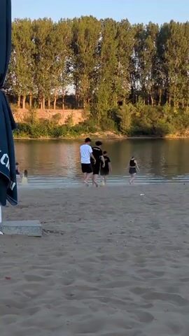 A group of young people are playing soccer on a sandy beach next to a body of water. The sun is setting, casting long shadows. The water is calm, and trees line the opposite bank. A large blue umbrella is partially visible in the foreground. The people are mostly dressed in casual summer clothing. One person is kicking a soccer ball towards others who are running to intercept it.