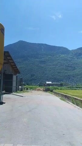 The video begins with a view of a paved area in front of a barn. A large yellow cylindrical object is partially visible on the left side of the frame. Beyond the paved area is a grassy field bordered by a concrete structure. In the distance, a mountain range covered in lush green trees dominates the background under a clear blue sky with a few scattered white clouds. The camera pans slightly to the right, revealing more of the field and the mountain range. The overall impression is a rural landscape on a sunny day.