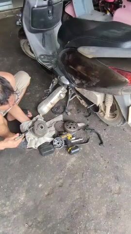 A man is kneeling on the floor in front of a disassembled scooter. He is wearing glasses and shorts. The scooter is black and partially disassembled, with various parts scattered on the ground, including a clutch and a drive face. The floor is concrete and appears dirty and oily. The man is holding a tool and working on the clutch assembly. The video is shot from a high angle, looking down at the scene.