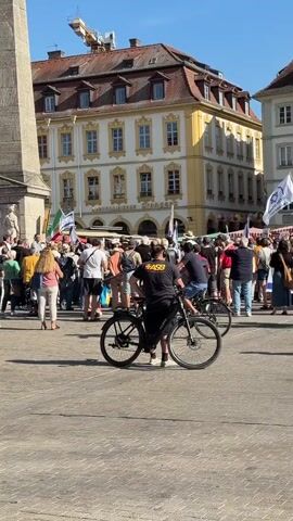 The video shows a crowded outdoor square on a sunny day. In the foreground, a black bicycle is parked. Behind the bicycle, a group of people are gathered, many holding flags. To the left, a tall stone obelisk is visible. In the background, there are buildings with red roofs and yellow facades. A construction crane is visible in the distance above the buildings. The people appear to be participating in an event or demonstration, with some interacting with each other and others observing. The atmosphere seems calm and the lighting suggests it is daytime.