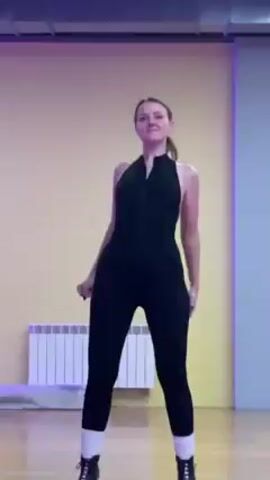 A woman in a black sleeveless jumpsuit is dancing in a studio. She is facing the camera and touching her chest. She has brown hair pulled back in a ponytail. The studio has a light yellow wall, a radiator, and a blue-grey column on the right. There is purple light at the top of the frame. The woman is smiling and bends backward with her arms out to the side, then places her hands behind her head.