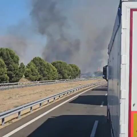 The video captures a road scene with a large white truck on the right side of the frame. The road appears to be a highway with multiple lanes and a guardrail. To the left of the road, there is a dry, grassy area with scattered trees and a large plume of dark smoke rising into the sky in the distance. The smoke suggests a fire is occurring, and some red flames are visible within the smoke. The sky is clear and blue, indicating daylight. The truck has a red stripe along its side and a yellow warning sign that reads "VEICULO LONGO." The overall environment appears to be outdoors, likely in a rural or semi-rural setting, affected by a fire.