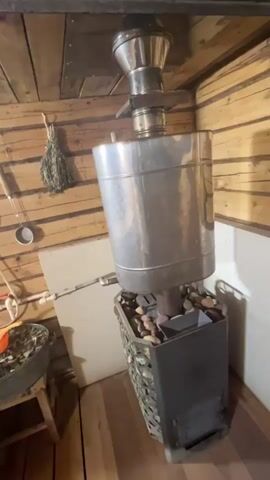The video showcases the interior of a sauna. A wood-burning stove with a stone basket and a water tank attached to a chimney sits in the corner of the room. The walls and ceiling are made of light-colored wood planks. A shower unit is mounted on the wall next to a bucket filled with green leaves. A small orange scoop rests on top of the leaves. Wooden benches line the side of the room. The video pans across the sauna, highlighting the different elements of the space.