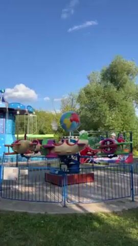 Here's a concise description of the video:

The video shows a children's airplane ride in an amusement park. The ride features several small, brightly colored airplanes attached to a central rotating platform. A globe sits atop the central structure. The ride is surrounded by a blue metal fence. There's a blue structure with stars next to it, and trees are in the background under a blue sky with a few clouds. The ride is not in motion.
