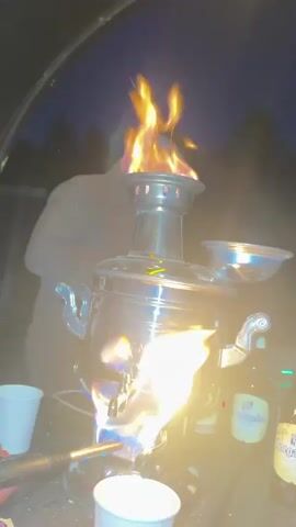 A samovar is being lit with a torch. The samovar is made of metal and has a round base and a tall, cylindrical body. It is sitting on a wooden table. There are several beer bottles and paper cups on the table. The background is dark, suggesting that it is nighttime. The flames are bright and yellow and orange. The torch is held by a person whose hand is visible in the lower left corner of the frame. The person's arm is also visible. The samovar has a tap on the front, from which water or tea would be dispensed. The lid of the samovar is open, and flames are coming out of the top. A person is visible in the background, standing behind the samovar. Their face is not visible. The fire is intense and appears to be burning well. The overall impression is one of an outdoor gathering or camping trip where hot drinks are being prepared.