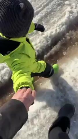 A person is holding the hand of a small child who is walking in slush. The child is wearing a neon green snowsuit and a dark knit hat. The slush appears to be dirty, with streaks of brown water mixed in with the melting snow. The person holding the child's hand is wearing a dark jacket and shoes. They appear to be navigating a snowy or icy path.