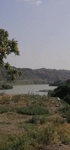 The video shows a rural landscape. The first frame displays a body of water surrounded by hills. A tree with green and yellow leaves is in the foreground. The second frame shows a truck parked on a dirt road next to a tree with similar foliage. The third and fourth frames depict a telephone pole in front of a brick building filled with hay bales. A large, barren mountain rises behind the building. The sky is clear and blue throughout the video.