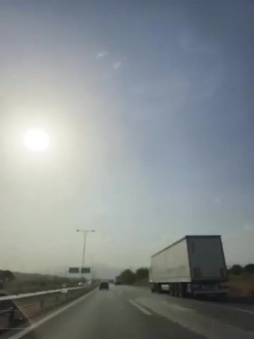 The video shows a POV shot from a moving vehicle on a highway under a bright, hazy sun. The sky is pale blue with some lens flares. To the right, a white semi-trailer truck is visible on the same highway. In the distance, a few other vehicles and mountains can be seen. There are streetlights and road signs along the side of the highway. The camera appears to be pointed slightly upwards towards the sky.