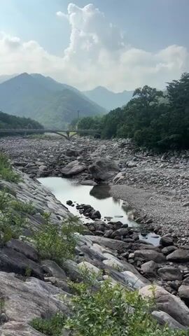 The video shows a rocky riverbed with a small stream flowing in one section. Lush green mountains and trees form the backdrop, with a clear blue sky and scattered clouds above. A stone bridge is visible in the distance. The camera pans across the scene, capturing the natural landscape. There are no people or animals visible in the video. The overall impression is of a serene and natural environment.