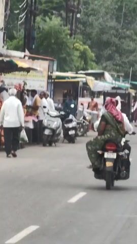 The video shows a street scene with several motorcycles and a bus. In the foreground, a person is riding a motorcycle from behind, with another person on the back. The person in the back is wearing a green sari and a pink headscarf. The motorcycle has a yellow license plate. To the left, there are some shops with people standing around. In the background, a green bus is visible with "MSRTC" written on it. The road is paved and appears to be in a rural or semi-urban area, with trees and vegetation visible in the background. The lighting suggests it is daytime. The overall atmosphere is that of a busy street with traffic and pedestrians.